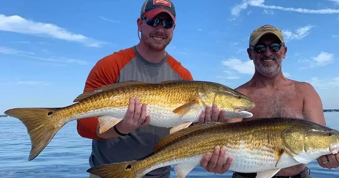 Two fishermen displaying redfish under blue sky on boat