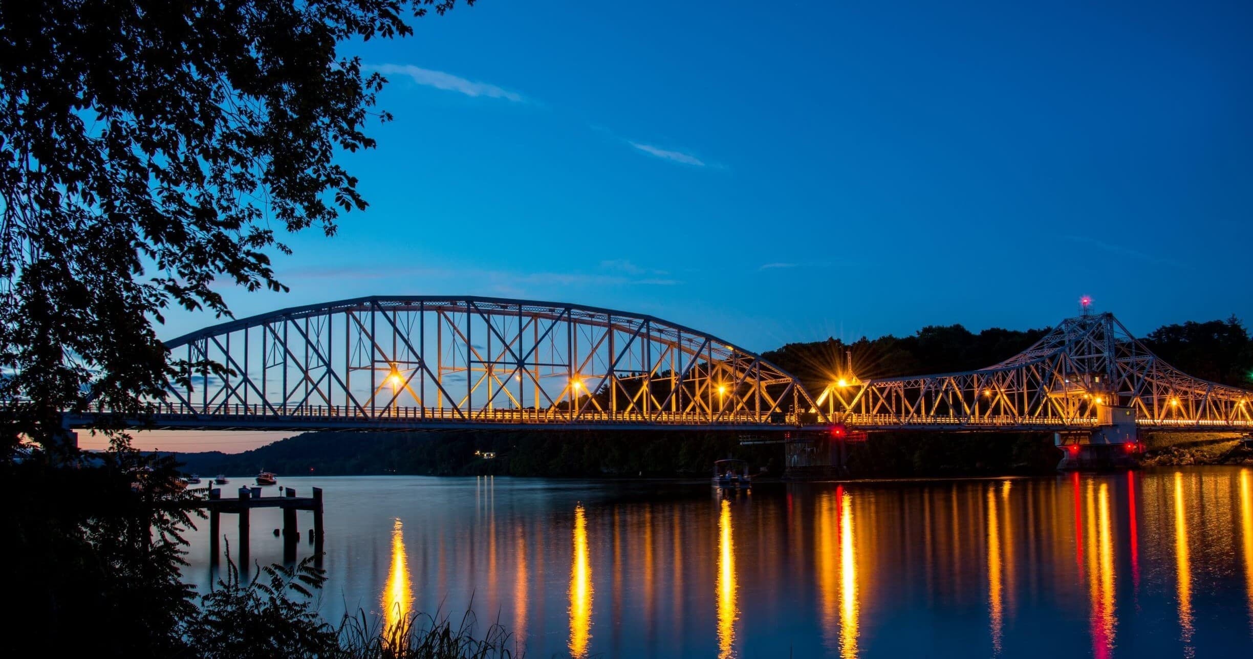 Illuminated truss bridge at dusk reflecting over calm riverfront