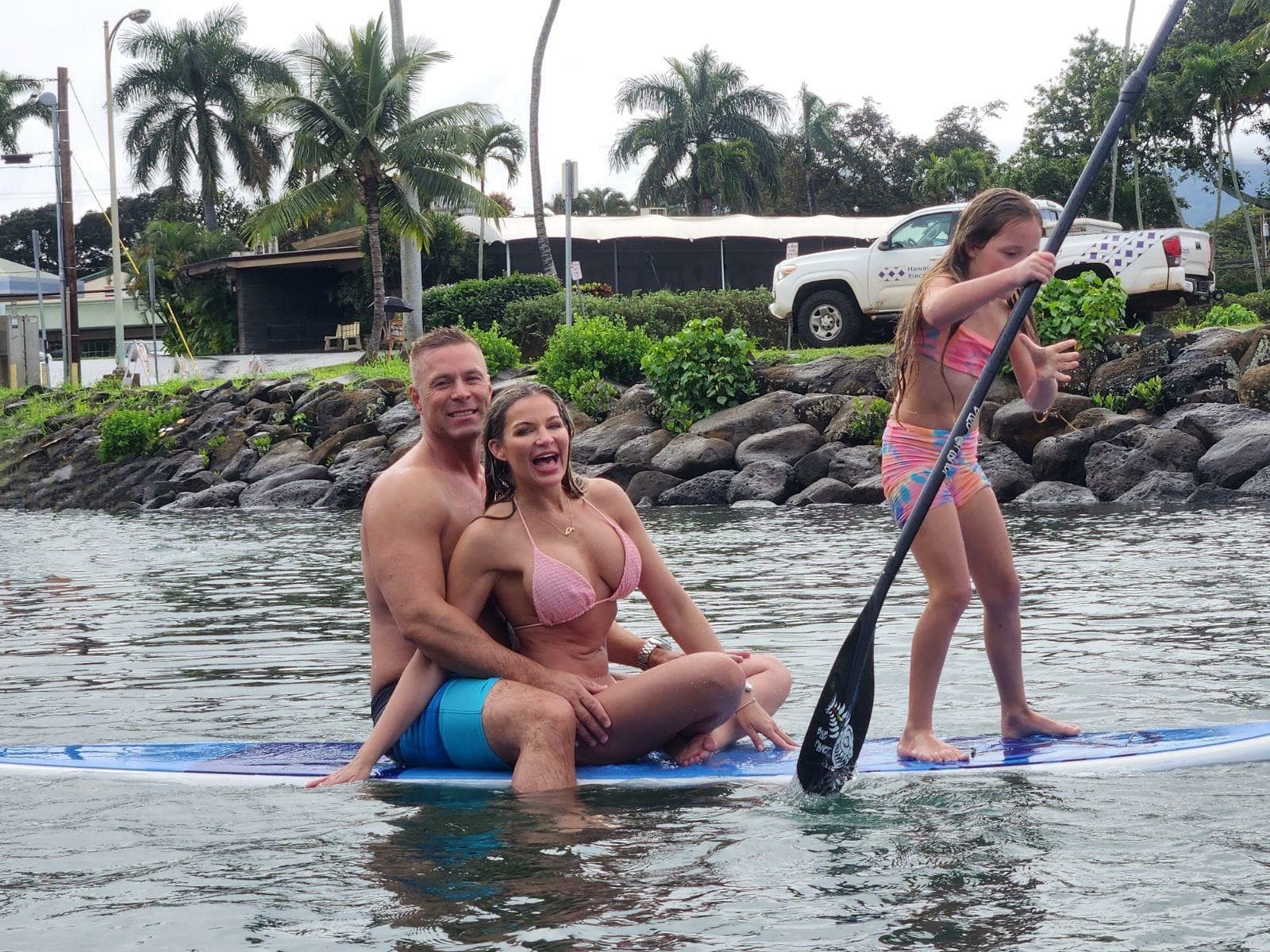 Family paddleboarding near palm-lined rocky shoreline, ocean