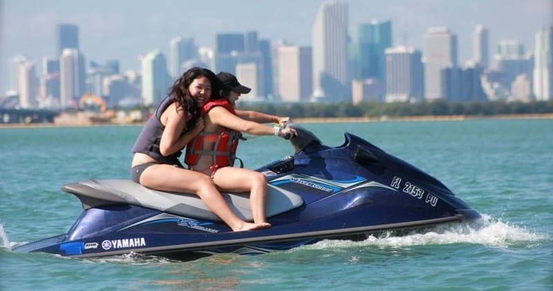Two riders on jet ski cruising with city skyline bay backdrop