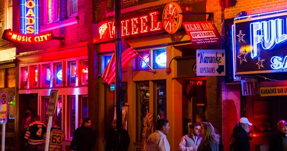 Pedestrians outside neon-lit bar entrances on Nashville music row