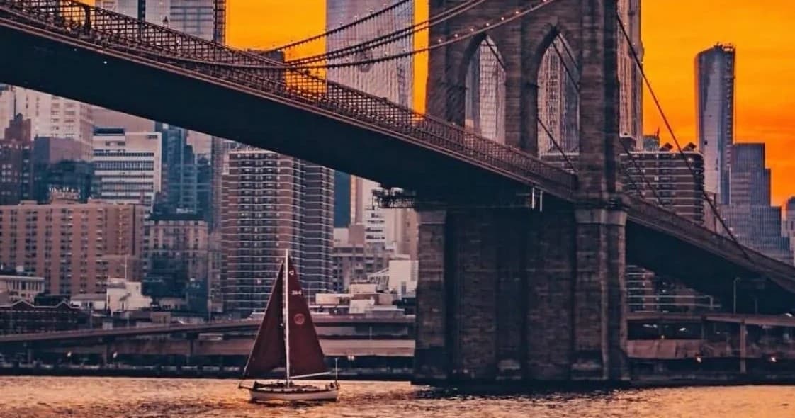 Sailboat under Brooklyn Bridge with urban waterfront views