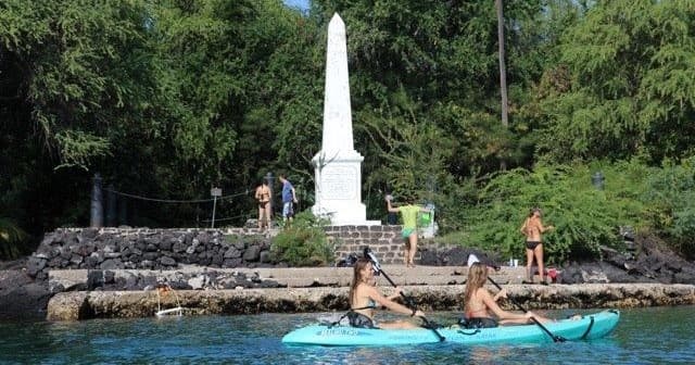 Two kayakers paddling near shoreline monument in tropical bay