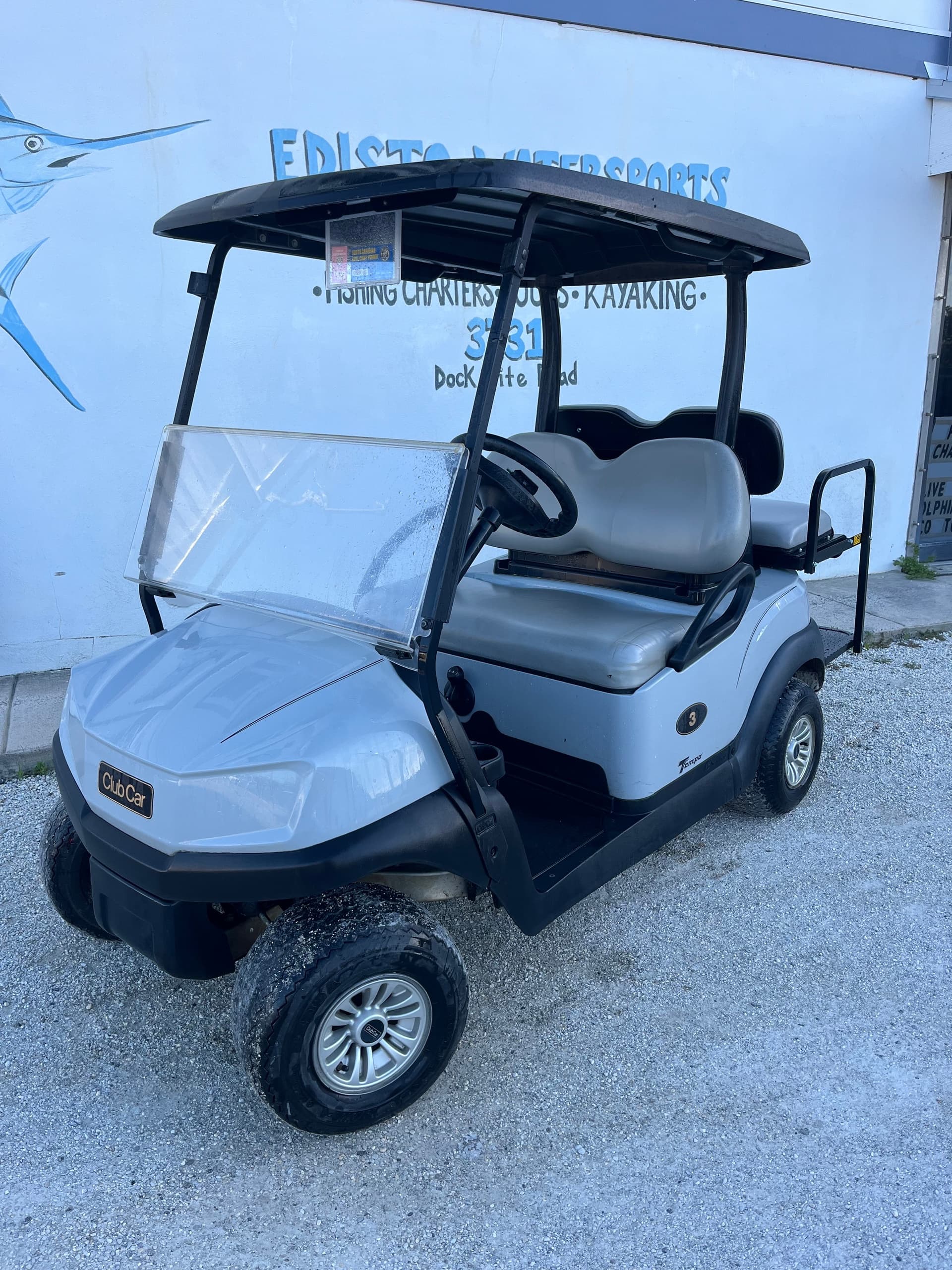 Light gray golf cart parked by waterfront shop on gravel lot