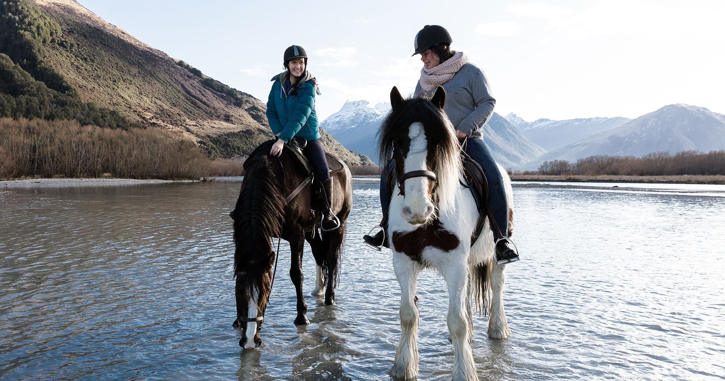 Two riders on horses wading in alpine lake with mountain views