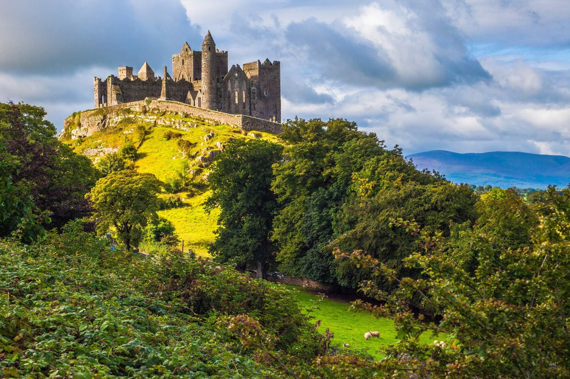 Historic hilltop castle overlooking green valley under dramatic skies