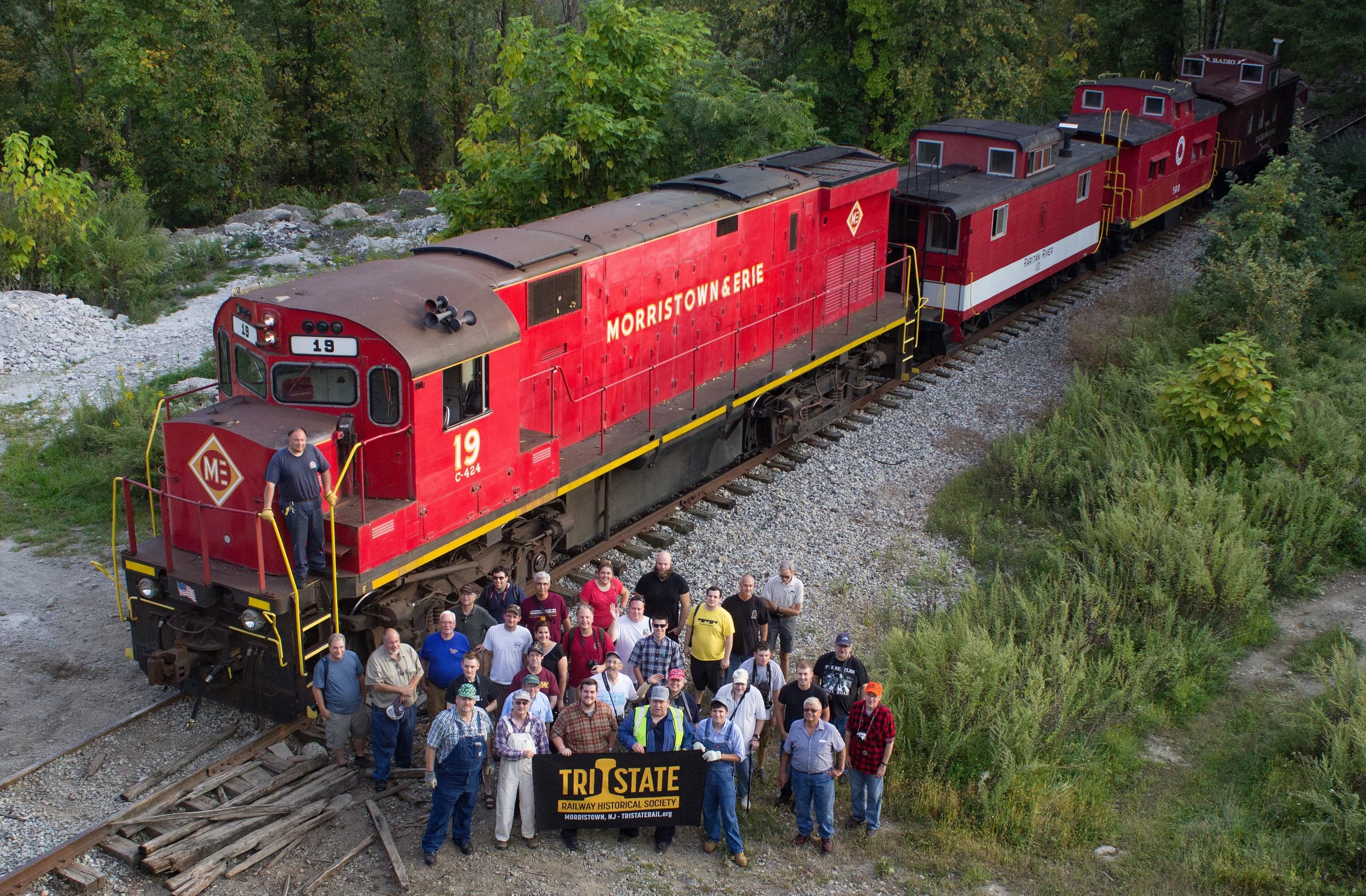 Red scenic train with crew and passengers posed by railroad tracks