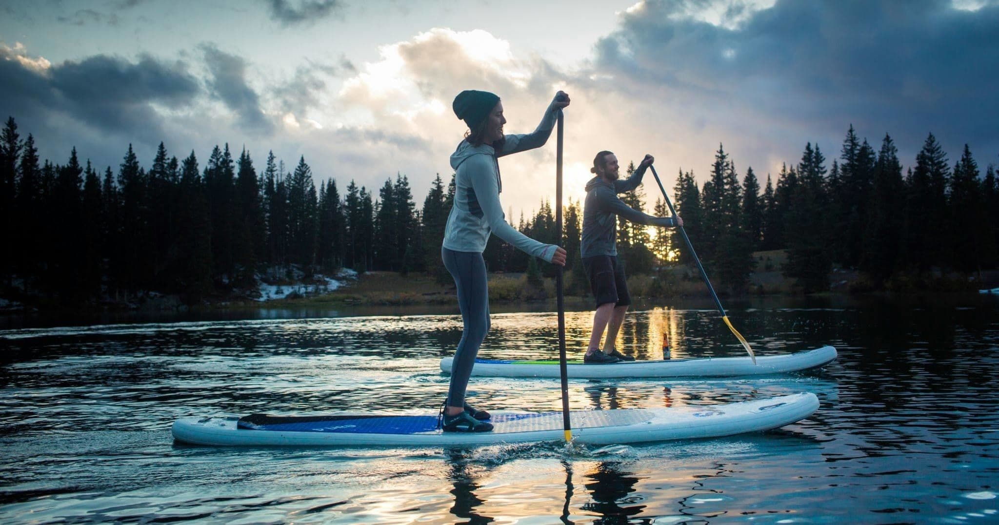 Two people paddleboarding at sunset on forested lake, silhouetted