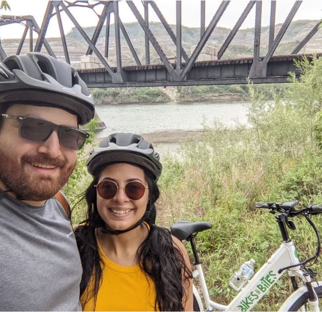 Couple smiling with e-bikes by river and railroad truss bridge