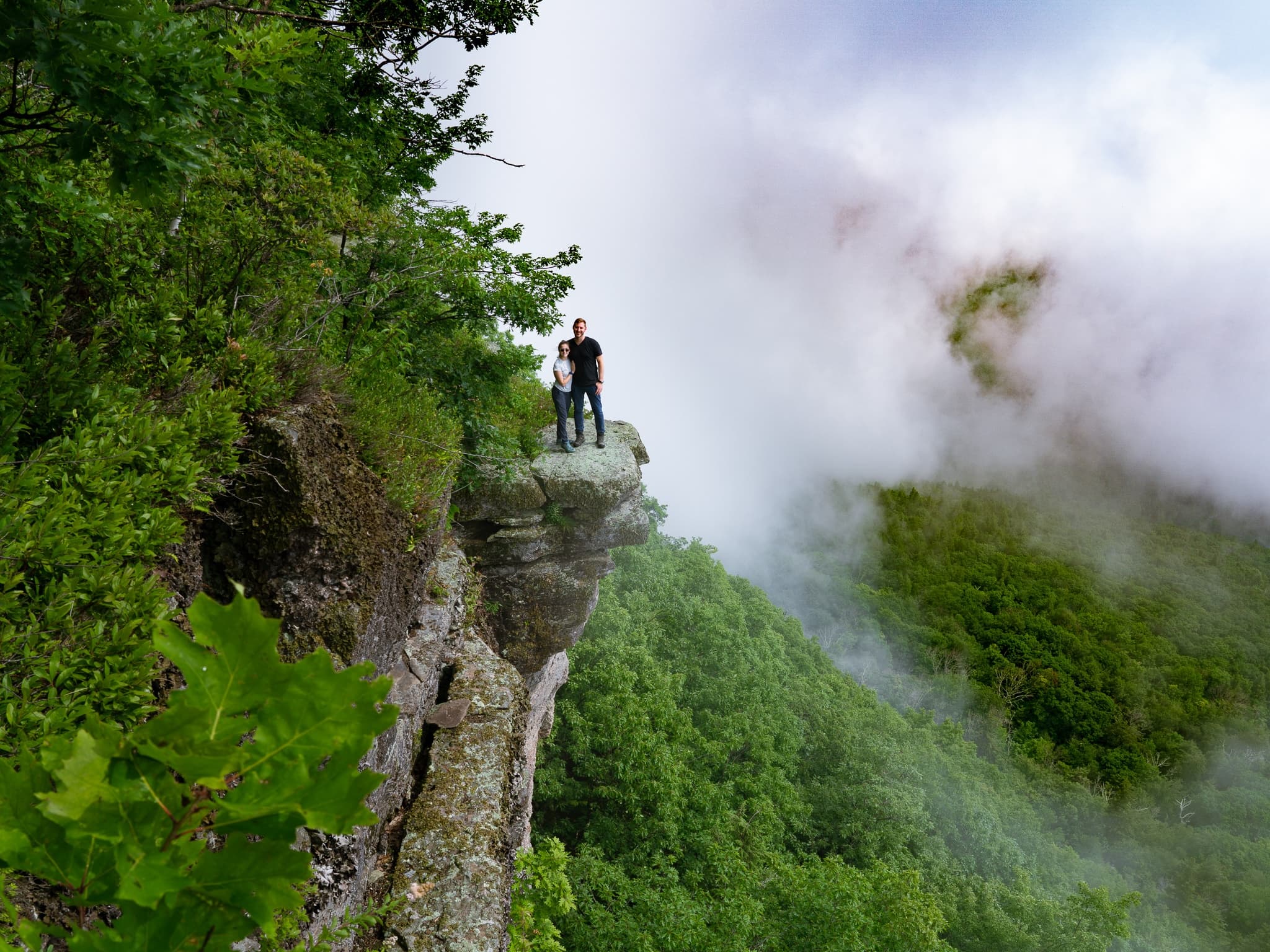 Couple standing on misty cliff edge above dramatic cloud valley
