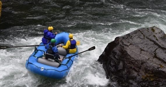 Rafters steering blue raft past large river boulder in rapids