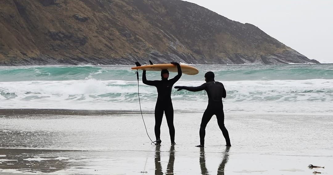 Two surfers carrying a longboard on reflective beach at low tide