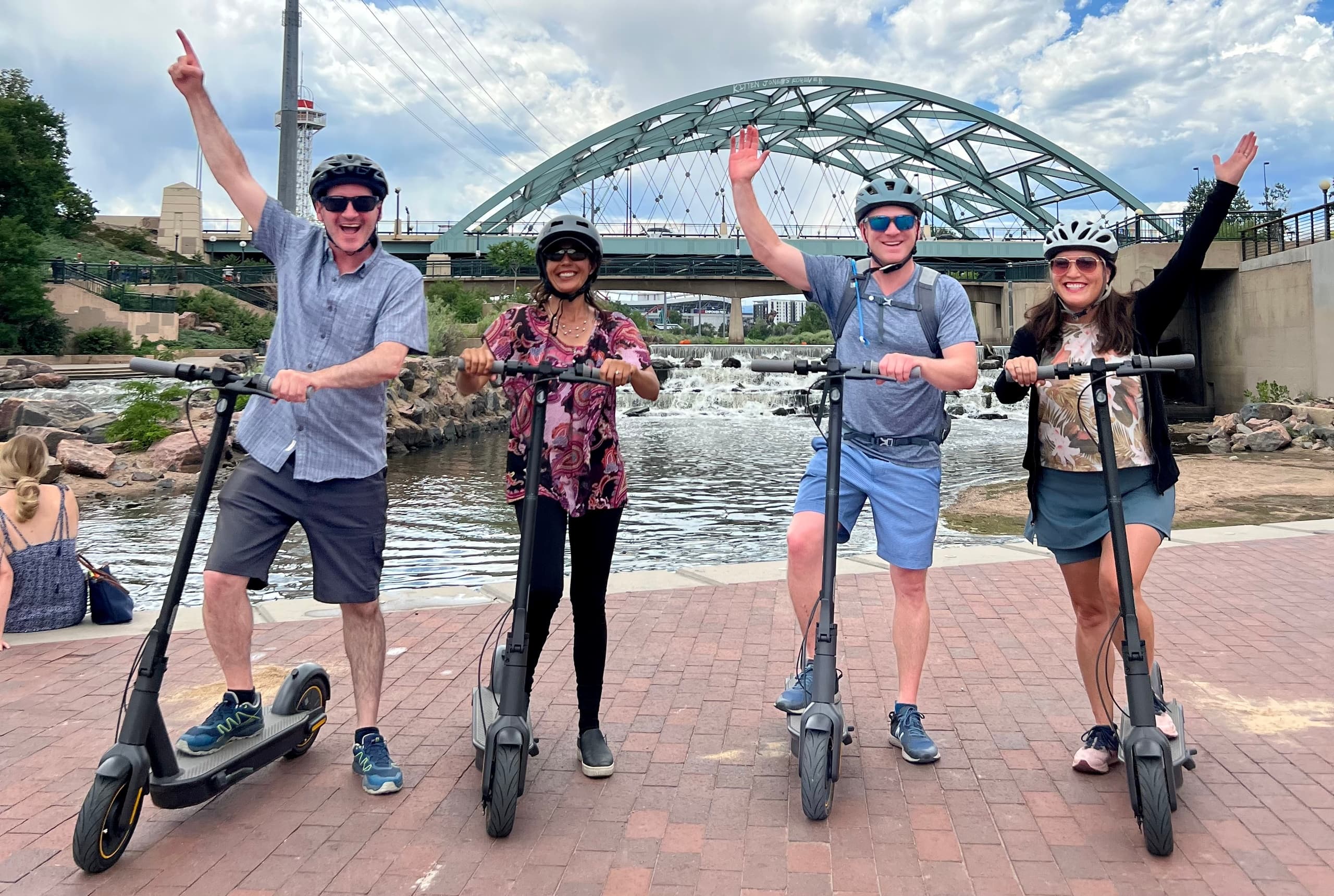 Group riding electric scooters along riverside under arched bridge