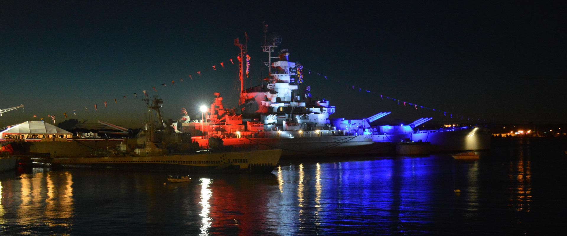 Illuminated battleship reflecting colorful lights on harbor at night