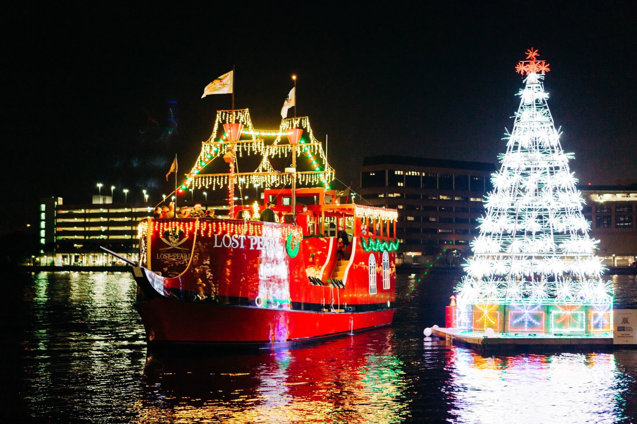 Festive pirate ship illuminated beside floating Christmas tree in harbor