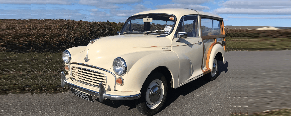 Cream Morris Minor estate driving along rugged coastal road Jersey