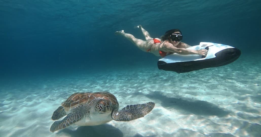 Snorkeler on reef scooter gliding past green sea turtle in clear shallow reef