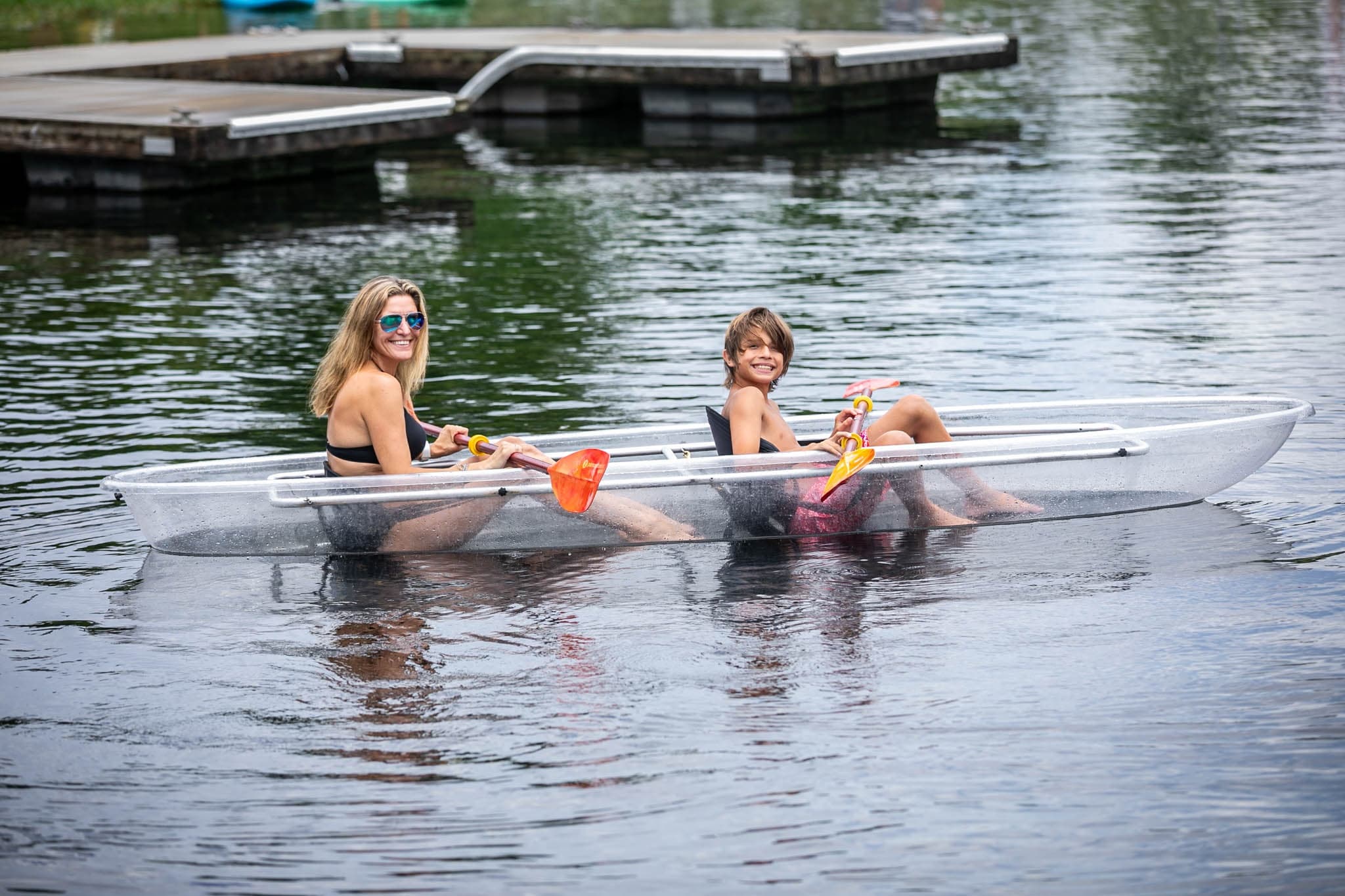 Transparent kayak with two paddlers near dock on calm lake