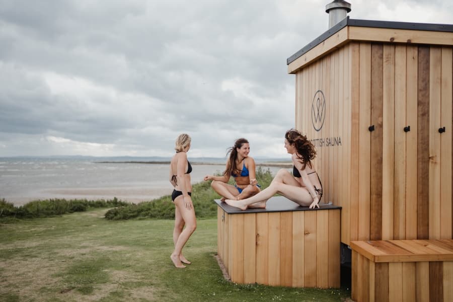 Women enjoying coastal ocean plunge beside wooden sauna on beach