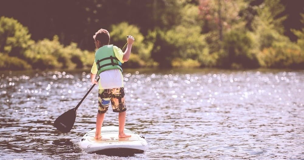 Child stand-up paddleboarding on calm lake near tree-lined shore