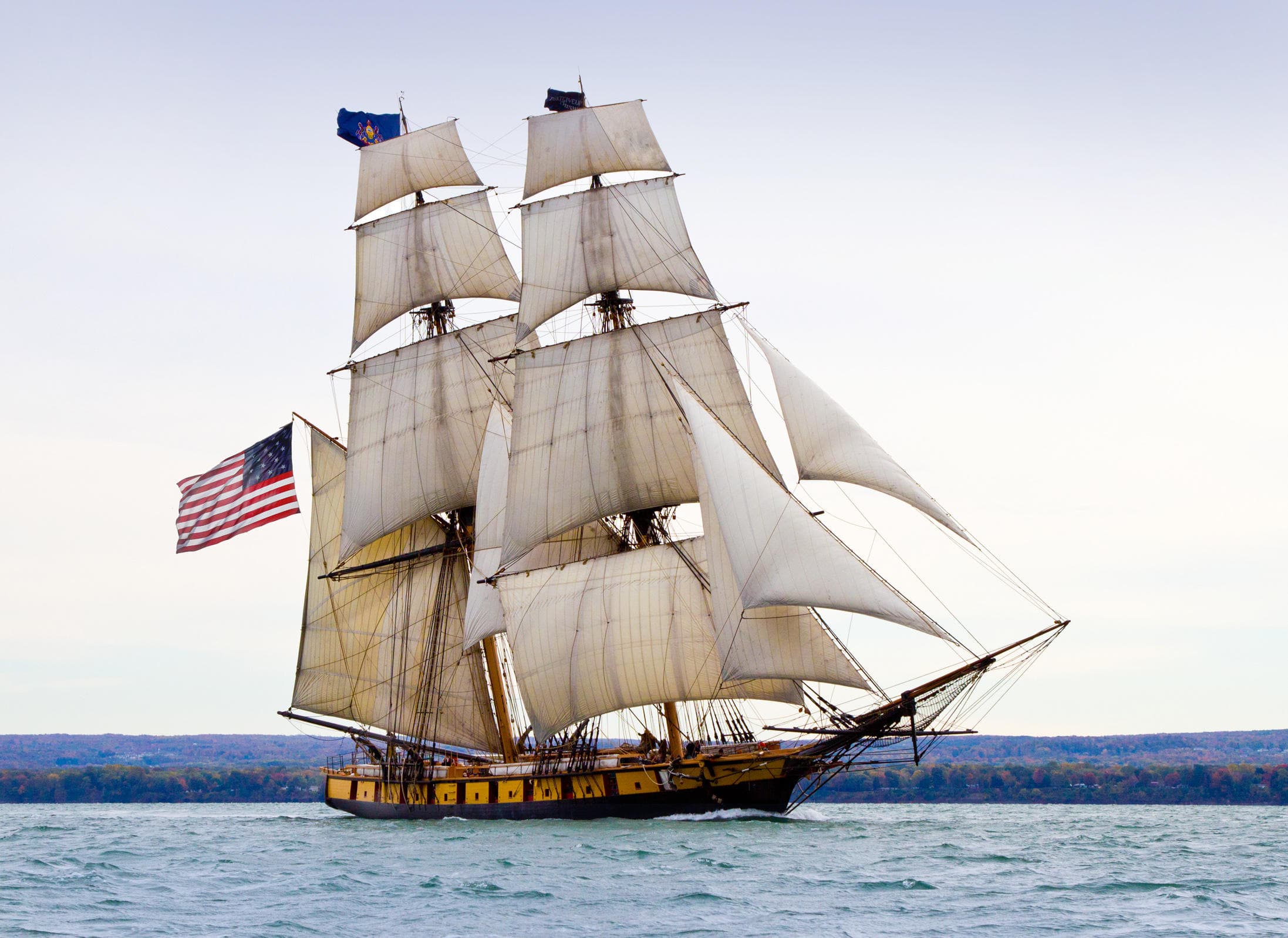 Three-masted tall ship sailing with American flag on open coastal waters
