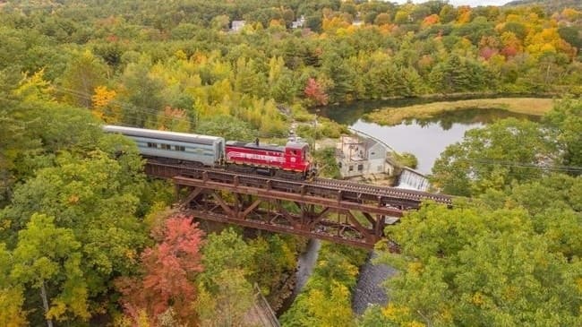 Aerial view of red train crossing a forested bridge in fall colors
