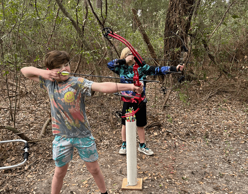 Kids practicing archery in wooded trail setting, aiming at target