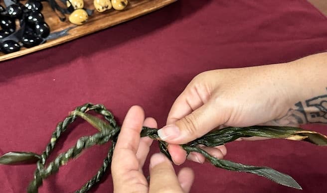 Hands braiding ti leaf lei at craft table during lei-making