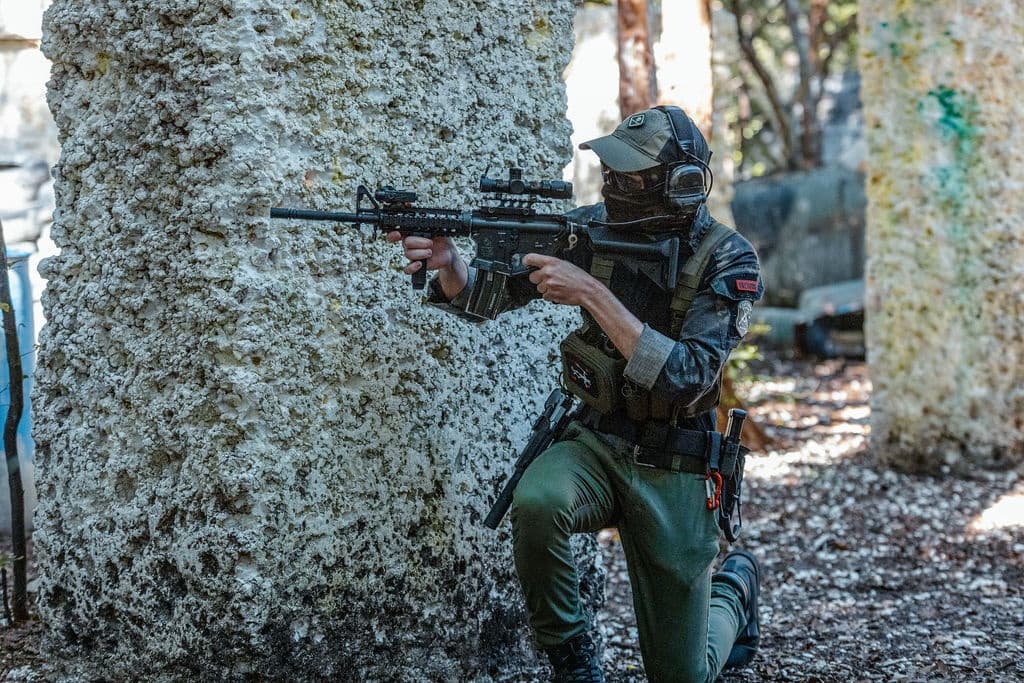 Player in woodland airsoft match aiming rifle behind cover