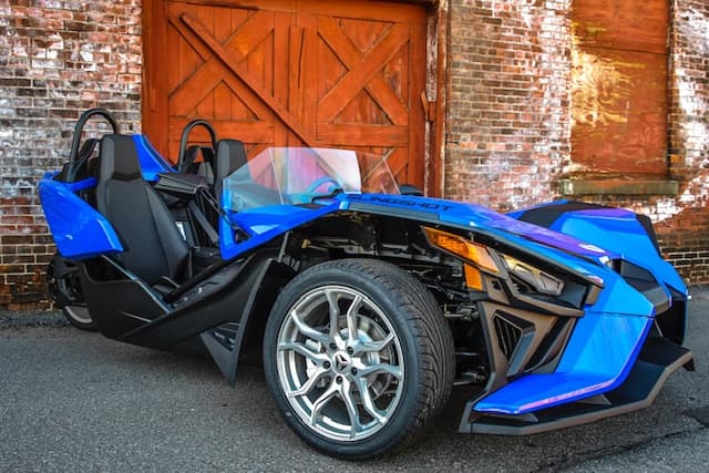 Blue Slingshot three-wheel roadster parked by brick warehouse