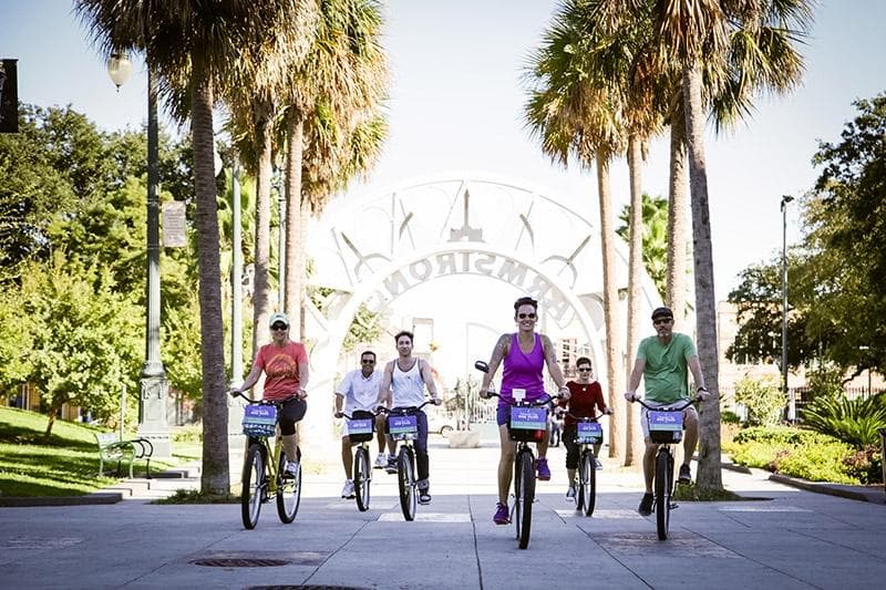 Bicyclists riding under palm trees at a waterfront park entrance in New Orleans