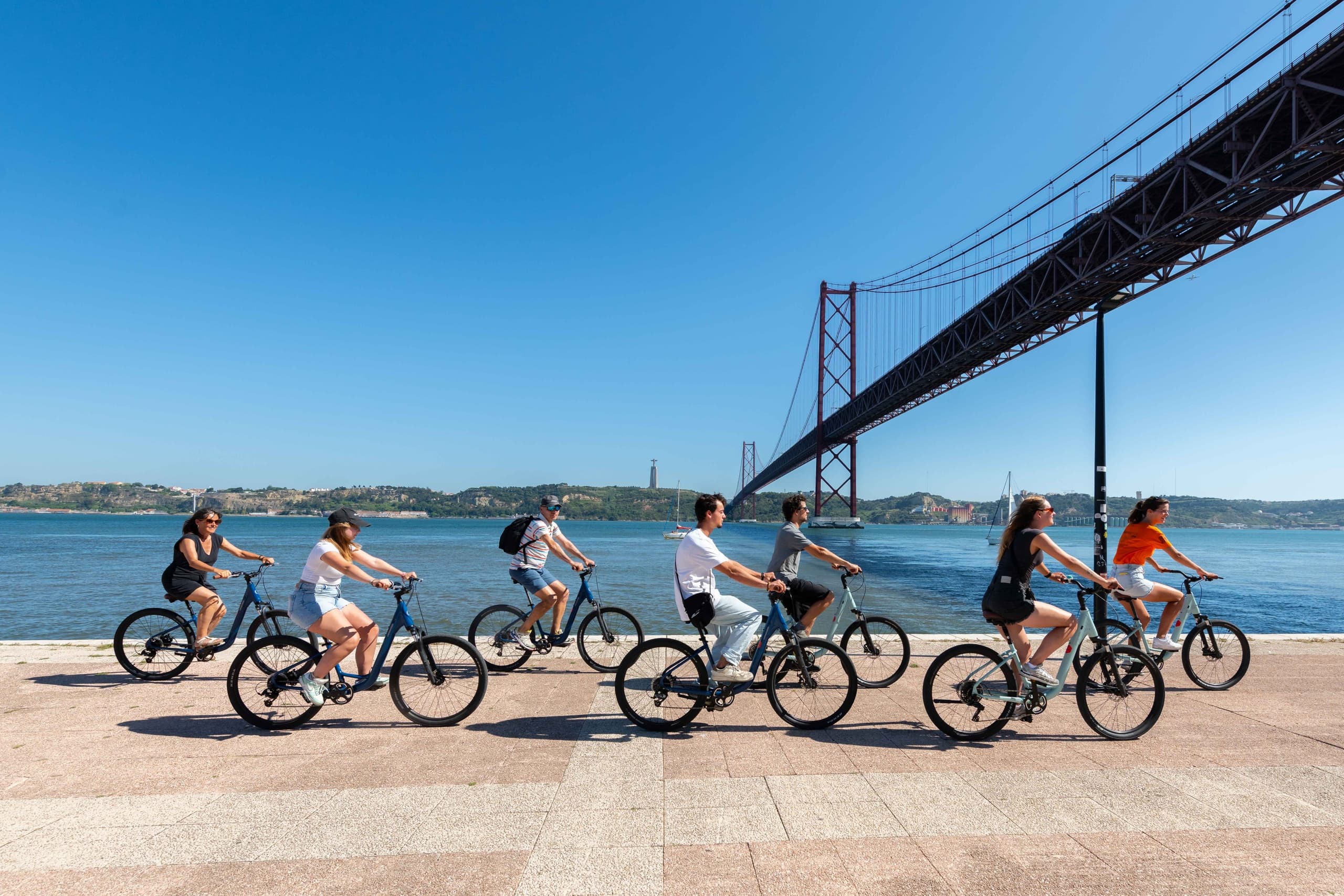 Cyclists riding along waterfront under Lisbon suspension bridge