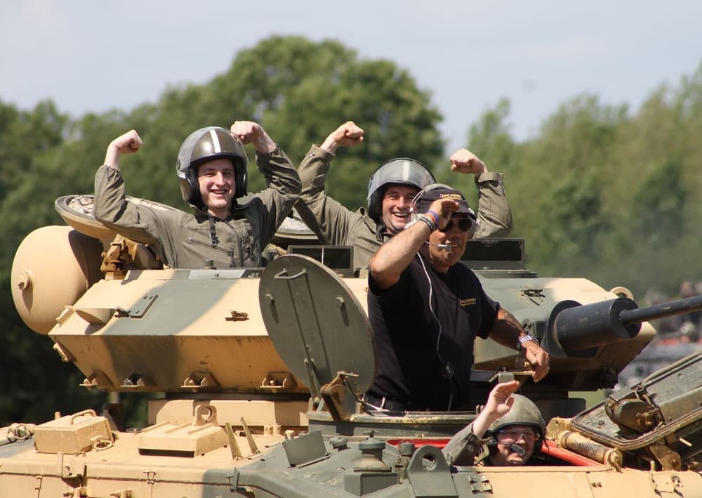 Participants cheering atop armored tank on sunny outdoor range
