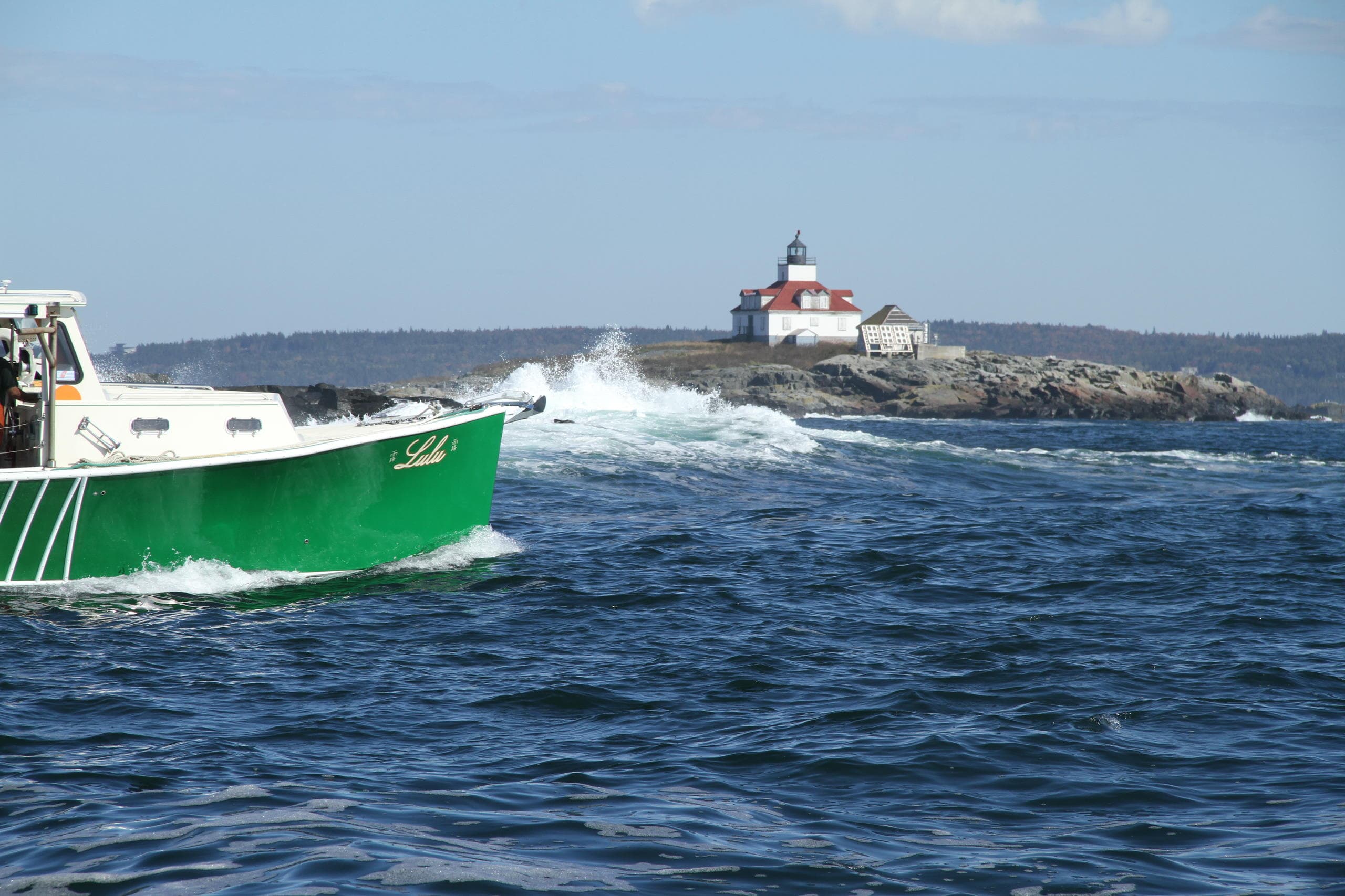Green tour boat speeding past coastal lighthouse and rocks