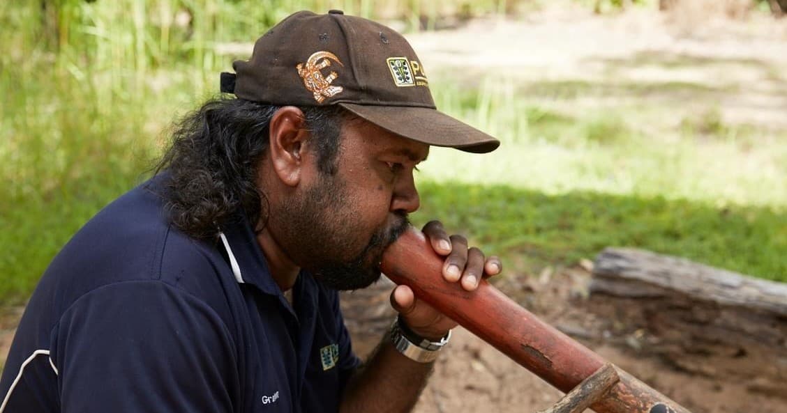 Indigenous man playing didgeridoo on grassy outdoor grounds