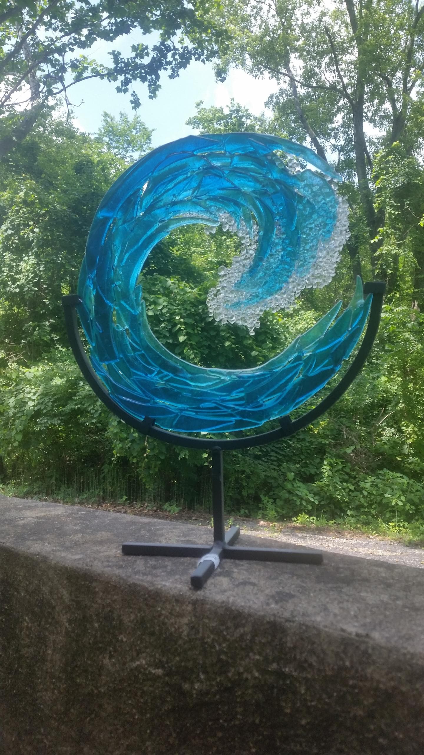 Blue glass wave sculpture displayed outdoors on park pedestal