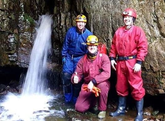 Cavers in wetsuits beside waterfall inside narrow canyon cave
