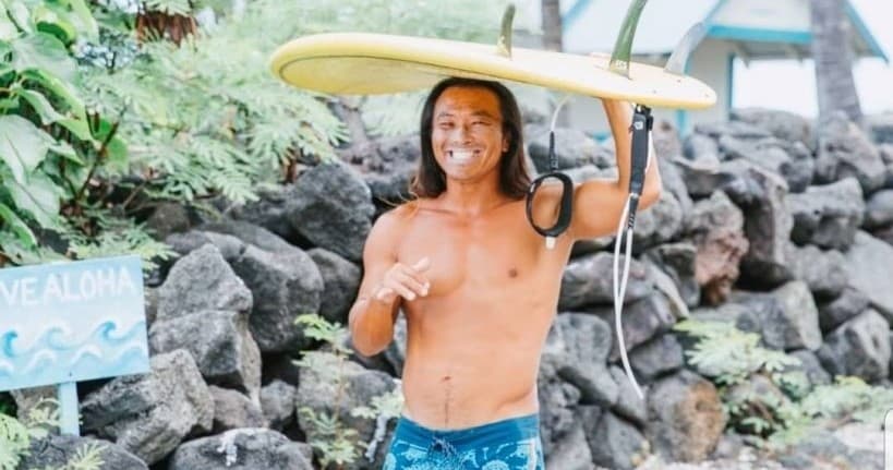 Smiling local surf instructor holding board on Kona lava rock shore