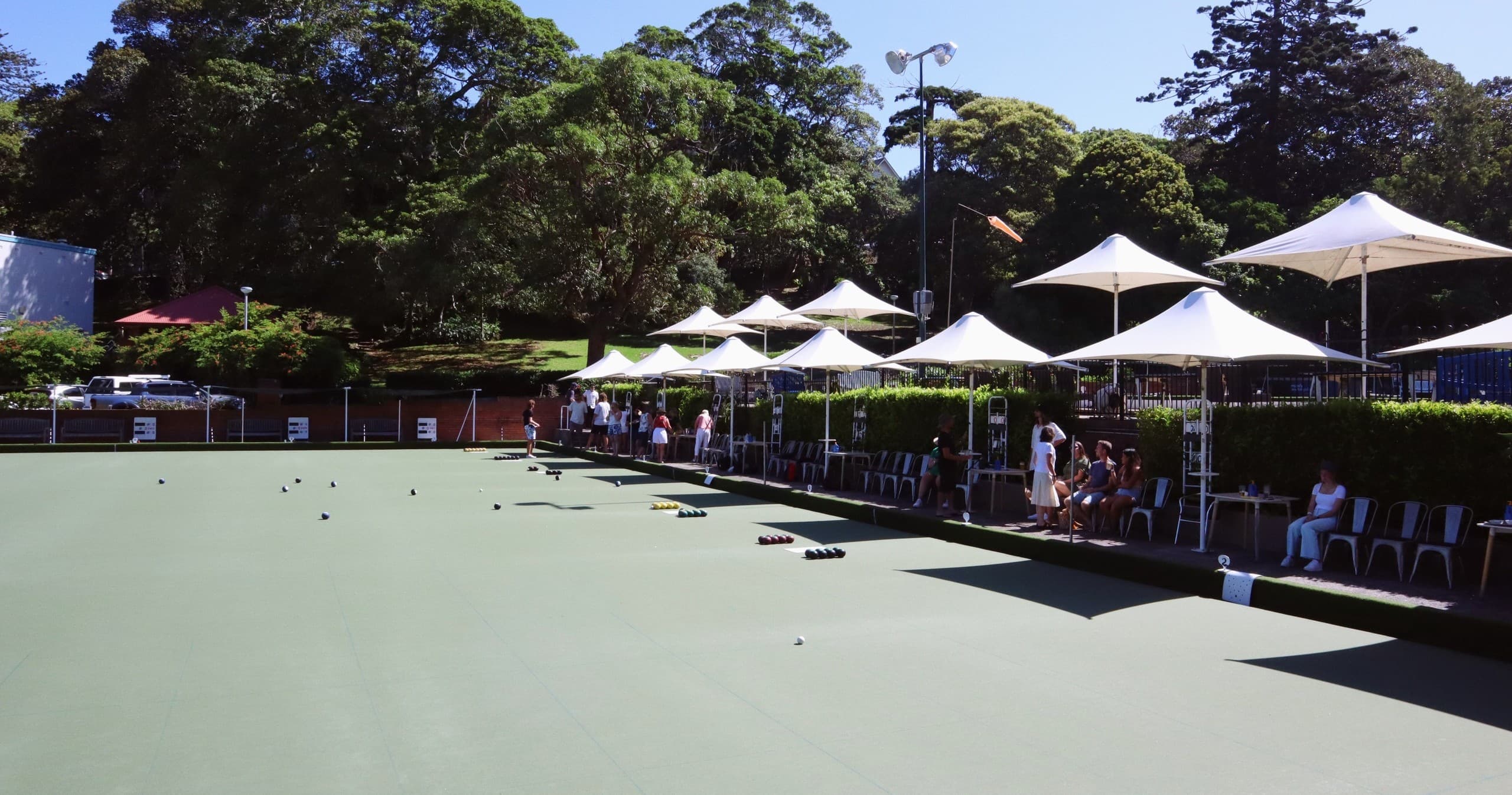 Wide lawn bowls green with players and shaded spectator seating