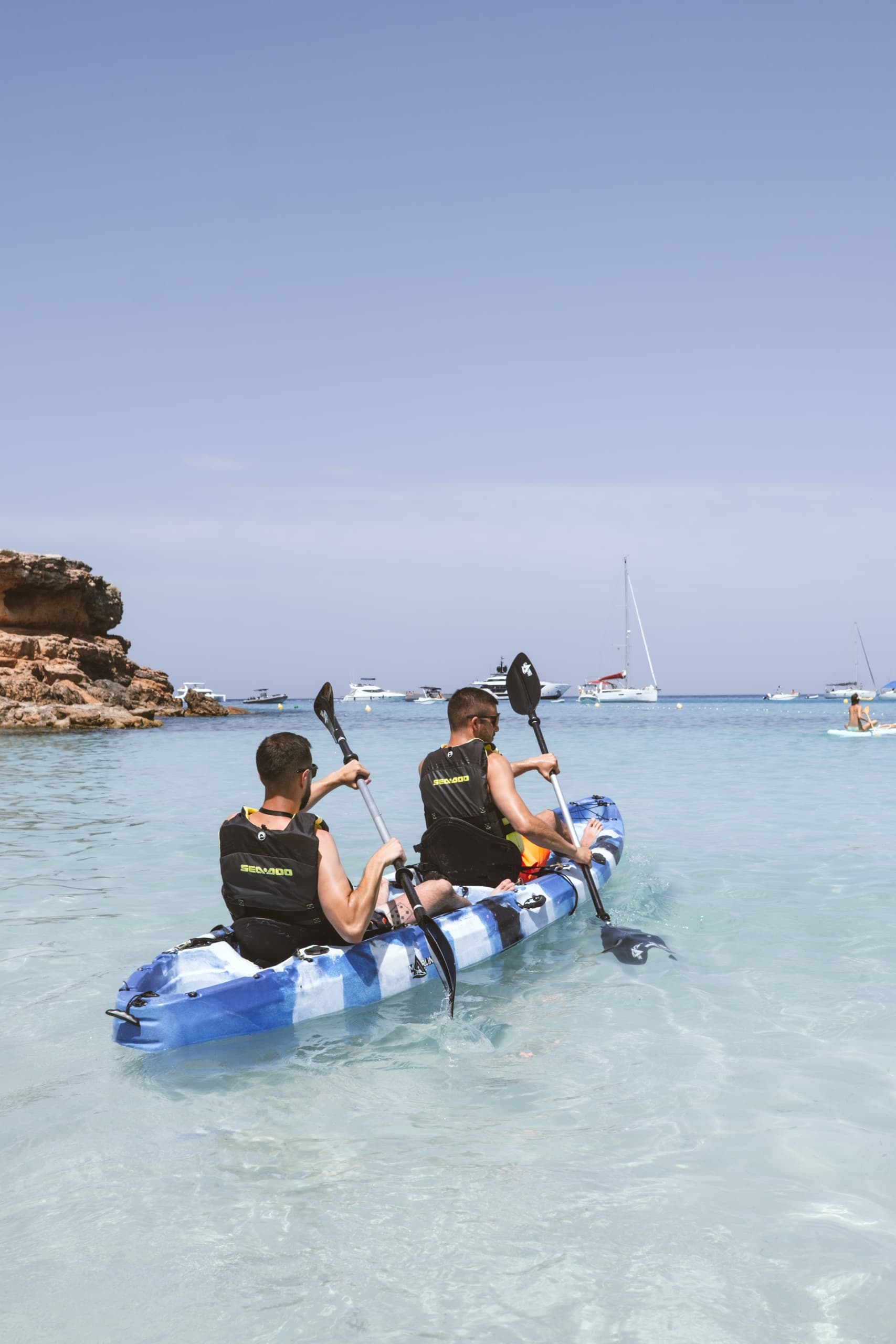 Two kayakers paddling in shallow clear bay near rocky beach