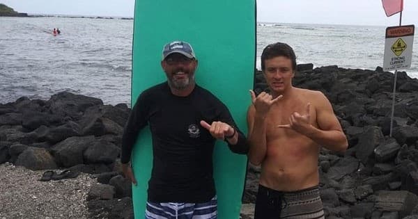 Instructor and student with surfboard on rocky shoreline by ocean