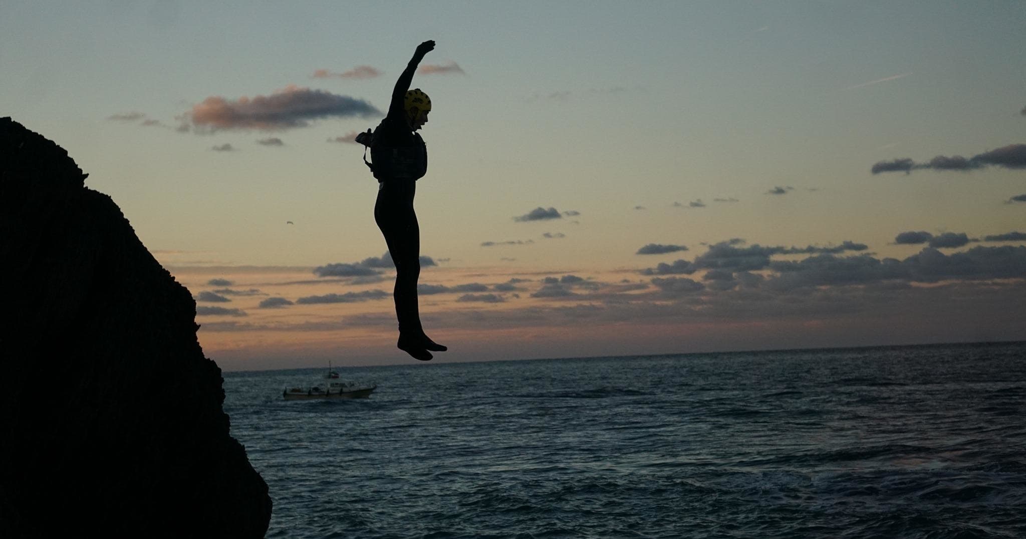 Silhouette cliff jumper leaping into ocean at sunset coastline