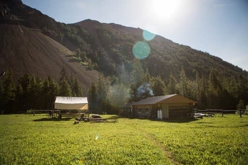 Sunlit cabins on grassy meadow below forested mountain ridge