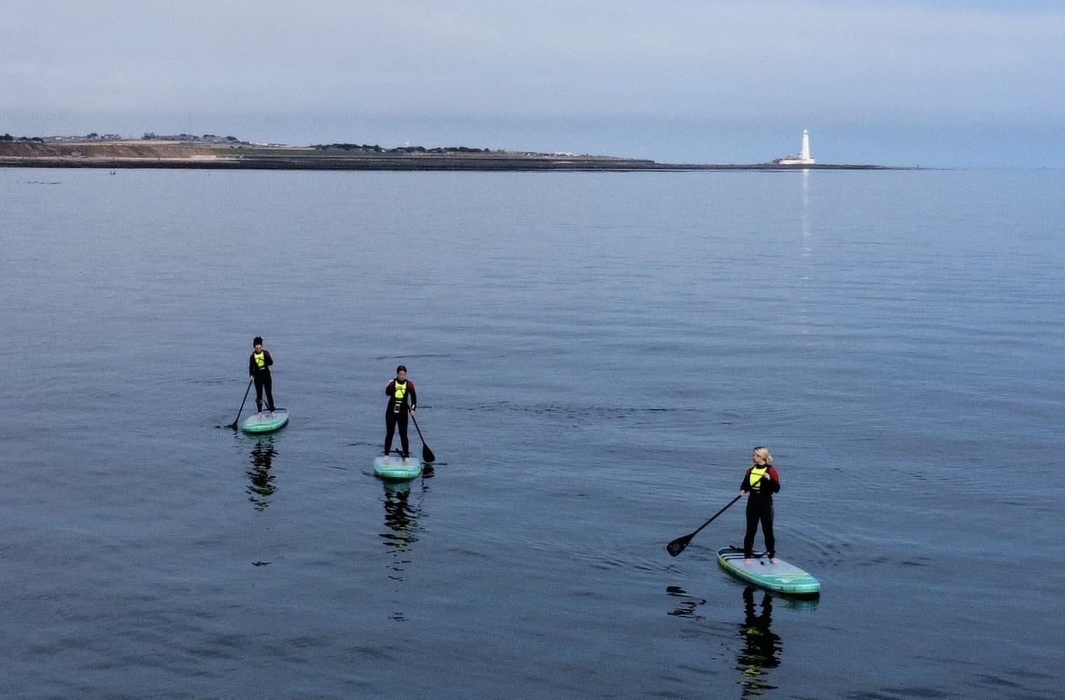Three paddleboarders gliding on calm sea toward distant lighthouse