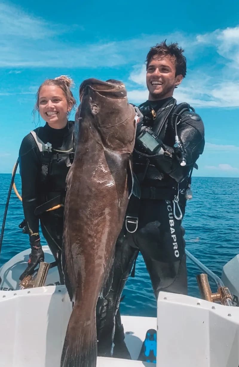 Two divers on boat holding large caught fish in open sea