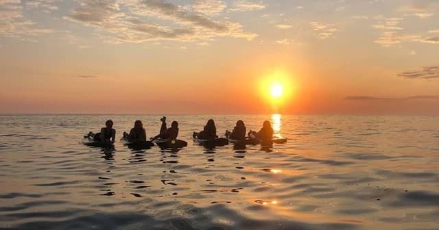 Group of surfers silhouetted at sunset on calm ocean horizon
