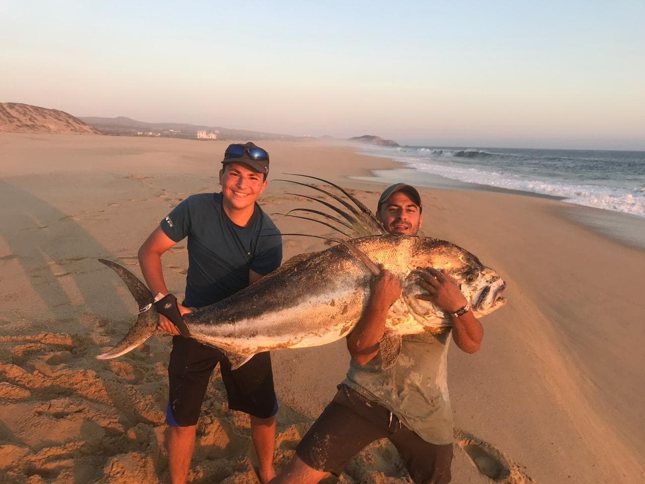 Two anglers holding giant fish on sandy Pacific beach at sunset
