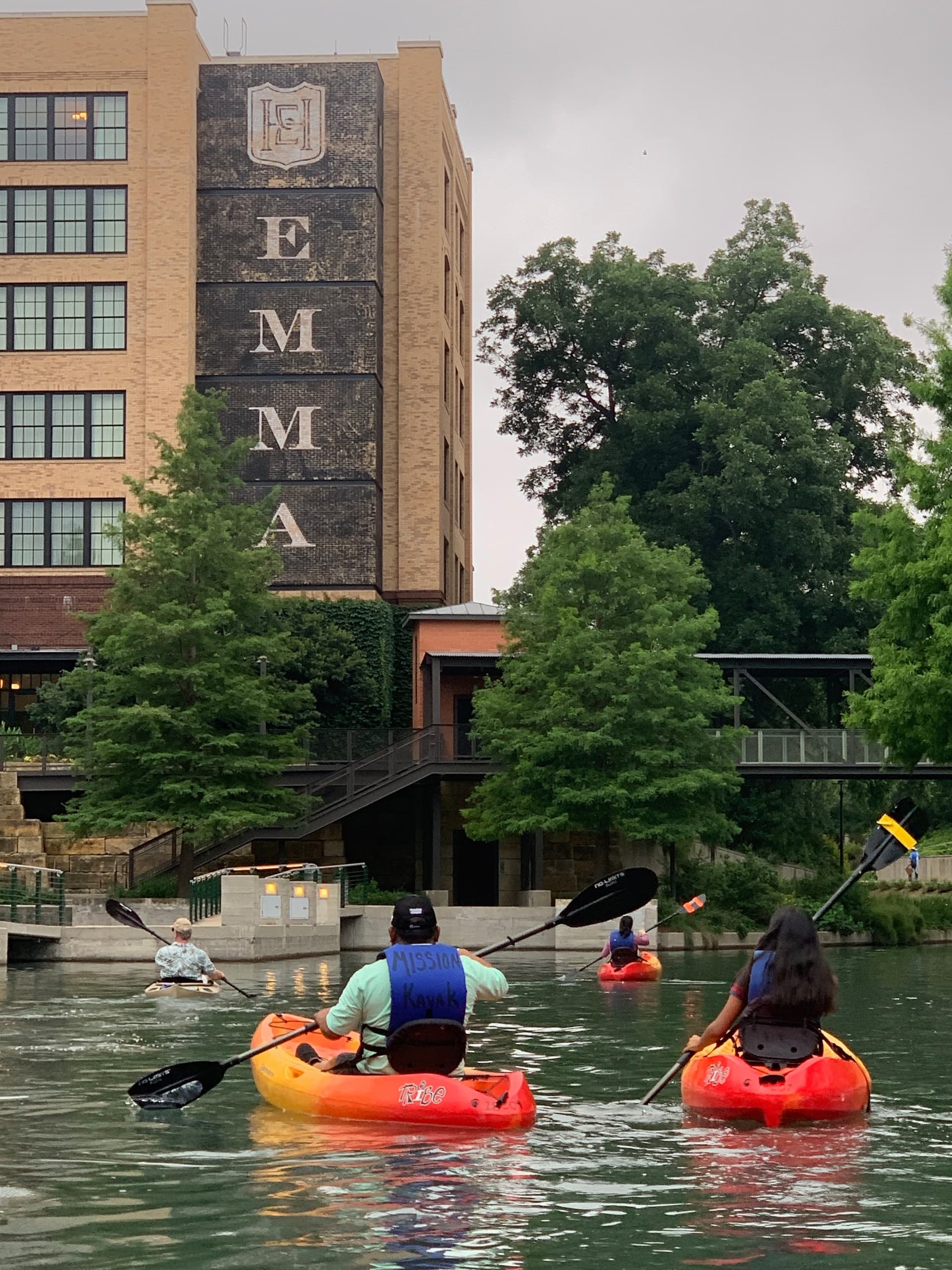 Two kayakers paddling past the EMMA building on San Antonio River