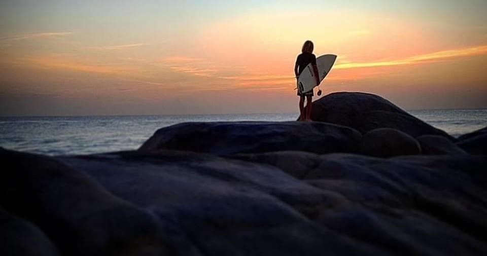 Surfer silhouette holding board at sunset on rocky beach