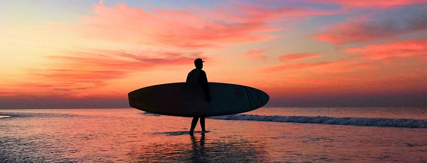 Surfer carrying board at sunset, coastal surf silhouette on beach